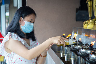 An asian woman in a white dress wearing a face mask is pouring oil into a lamp to make a wish