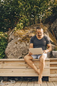 Full length of mature man using laptop while sitting on patio against trees