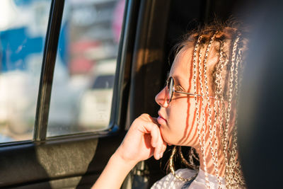 A pensive teenager girl with afro-braids and sunglasses looks out the window while 