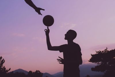 Cropped hand of man gesturing while friend playing with ball against sky during sunset
