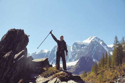 Man with arms raised holding hiking pole while standing on rock against mountains and sky