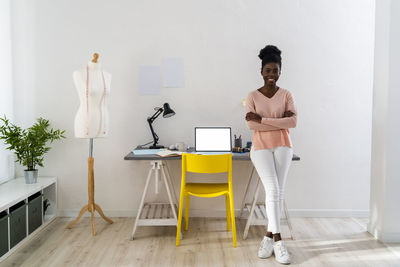 Confident fashion stylist with arms crossed smiling while leaning on table at home