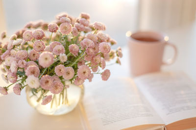 Small pink flowers bouquet in glass vase with blurred background of pink cup of tea and opened book 
