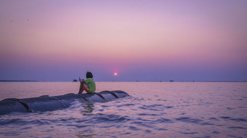 Woman sitting on pipe in sea against sky during sunset