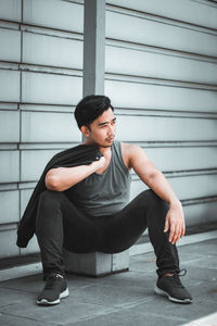 Young man looking away while sitting on footpath against wall