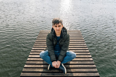 A man sits relaxed on a wooden pier on the lake, cross-legged in jeans and a jacket in autumn