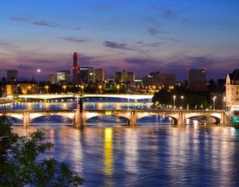 Illuminated bridge over river by buildings against sky in city