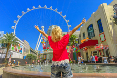 Rear view of woman with arms raised standing by fountain and buildings in city