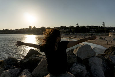 Rear view of woman sitting on rock by sea against sky