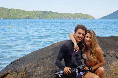 Portrait of smiling couple sitting on rock by sea