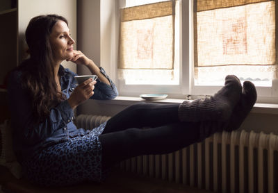 Daydreaming woman with feet on the heater drinking cup of tea while looking through window