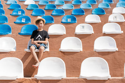 Serious boy in summer clothes sitting on one of chair on stadium and looking to camera