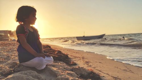 Girl looking away while kneeling at beach against clear sky during sunset