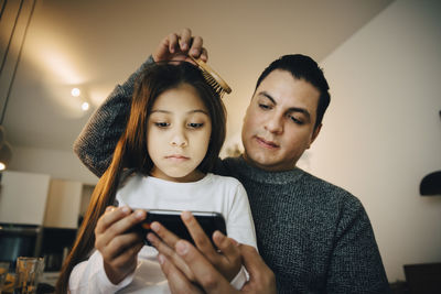 Low angle view of father combing daughter's hair while girl using mobile phone at home