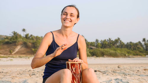 Portrait of smiling young woman sitting on beach against clear sky