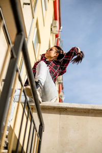 Low angle view of woman tying hair while sitting in balcony against sky