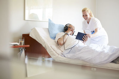 Happy female nurse assisting senior man in using digital tablet on hospital bed