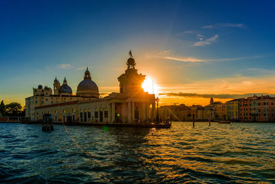 Church by canal and buildings against bright sun in sky during sunset