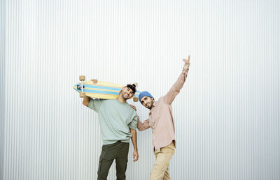 Two men standing with longboard in front of white wall