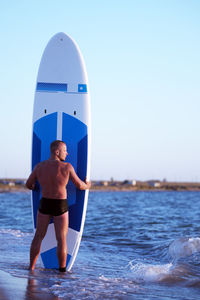 Rear view of shirtless male surfer holding surfboard at beach against clear sky