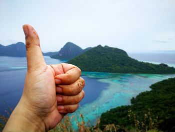 Close-up of cropped hand showing thumbs up by sea against sky