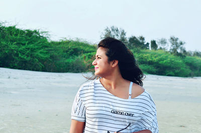 Beautiful young woman looking away standing against sky at beach