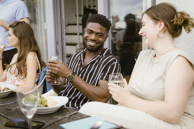 Portrait of smiling young woman having drinks with female friend sitting at dining table during dinner party in balcony