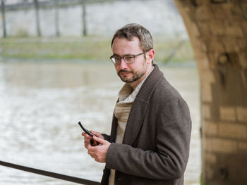 Man holding mobile phone while standing against lake