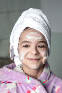 A girl with a bath towel on her head and cleansing foam applied to her face.