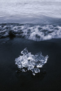 From above of chunk of shiny ice on black sandy beach washing by wavy ocean on cloudy day in iceland