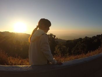 Side view of woman standing by retaining wall on mountain against sky during sunset