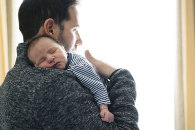 Father with sleeping baby boy looking away while standing by window