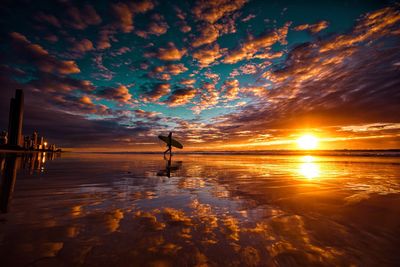 Silhouette man with surfboard walking at beach with clouds reflection at sunset