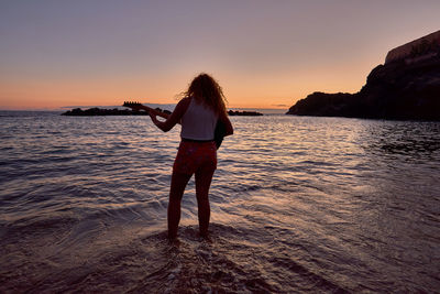 Rear view of woman standing on beach against sky during sunset