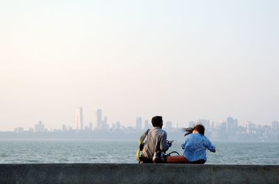 Rear view of man and woman sitting by sea at marine drive against clear sky