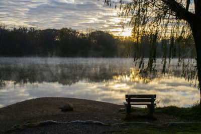 Bench by lake against sky during sunset