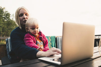 Mother using laptop while holding daughter at table in holiday villa