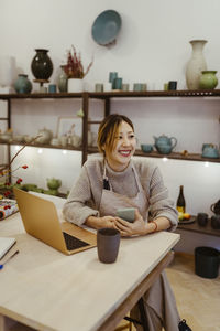 Happy female potter with laptop and smart phone sitting at workshop