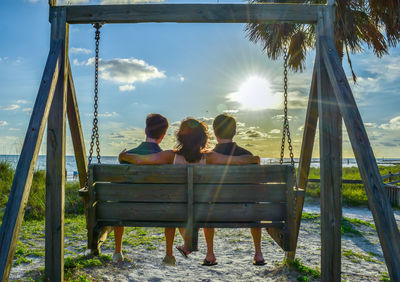 Rear view of friends sitting on swing at beach against sky