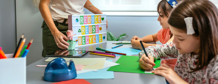 Teacher holding lightbox with text reduce, reuse, recycle while students drawing in classroom