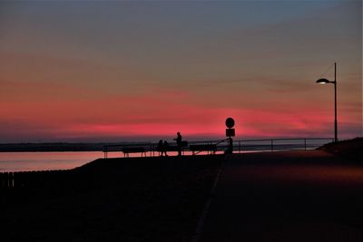 Silhouette people on street by sea against orange sky