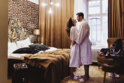 Side view of romantic couple embracing while standing by bed in hotel room