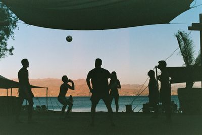 Silhouette people playing soccer on beach against sky