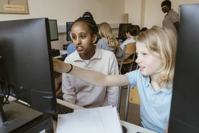 Girl pointing at computer monitor while sitting with female friend in classroom at school