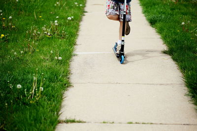 Low section of boy riding push scooter on footpath at grassy field