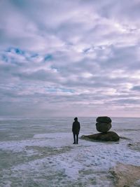 Rear view of man standing by rocks in sea against cloudy sky