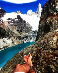 Low section of man resting on rock by river and snowcapped mountain against sky