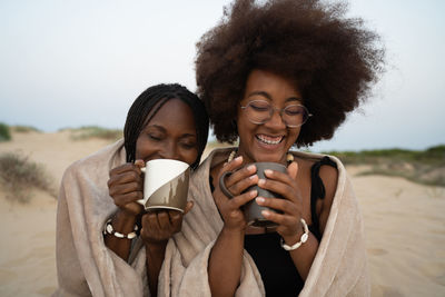 Happy young black female best friends with mugs of hot drinks sitting close wrapped in warm blanket and having fun during summer evening on sandy beach