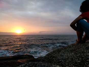 Woman sitting on rock by sea against sky during sunset