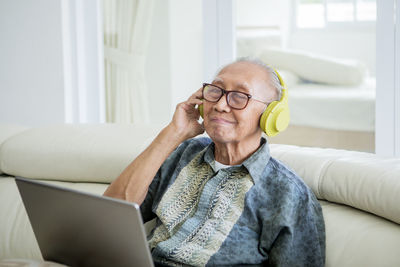 Smiling senior man with laptop wearing headphones while sitting on sofa at home